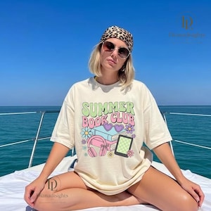 May include: Cream-colored t-shirt with the text "Summer Book Club" in pastel colors. The graphic includes books, headphones, flowers, and a tablet. The shirt is worn by a person on a boat.