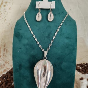 Peut inclure: Un ensemble de collier et boucles d'oreilles en argent. Le collier présente un grand pendentif vertical avec un design côtelé. Les boucles d'oreilles et le pendentif ont l'apparence d'un coquillage. Les bijoux sont exposés sur un buste en velours vert.