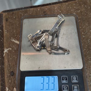 Peut inclure: Un collier en argent aux détails complexes est posé sur une balance numérique. La balance affiche un poids de 33,3 grammes. Le collier est composé de plusieurs segments liés avec un motif texturé.