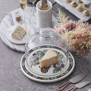 May include: A cheese dome with a glass lid, showcasing a wedge of cheese on a decorative plate with a butterfly and floral design. The plate is white with a black and red border. A marble serving tray and wine glasses are also visible.