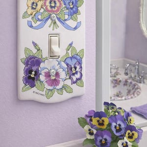 May include: White ceramic light switch cover decorated with colorful pansies. The floral design includes purple, yellow, and blue flowers with green leaves and a blue ribbon. A vase of pansies sits on the counter.