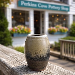 May include: A handcrafted ceramic vase with a cylindrical shape. The vase features a gradient of colors, transitioning from tan to black. The rim is white. The background shows a shop with the text "Perkins Cove Pottery Shop".