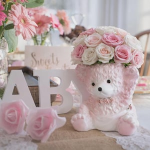 May include: A pink teddy bear-shaped vase filled with pink and white roses, surrounded by the word "BABY" in white letters and pink roses. Other flowers in vases are visible in the background. The word "Sweets" is also visible.