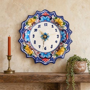 May include: A round wall clock with a scalloped edge, featuring a cream-colored face with dark blue numerals. The border is decorated with hand-painted floral patterns in vibrant shades of yellow, blue, red, and dark blue on a white background.