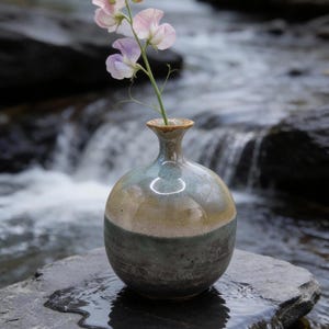May include: A small, round ceramic vase with a narrow neck and a flared rim. The vase features a two-tone glaze, with a glossy light tan upper section and a darker, mottled blue-green lower section. A single stem of pale pink sweet pea flowers is inside.