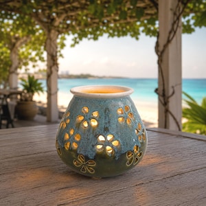 May include: A ceramic wax warmer with a rounded, bulbous shape featuring a two-tone glaze of teal and dark green. The exterior is decorated with cut-out flower patterns that allow warm, glowing light to shine through from the interior basin.
