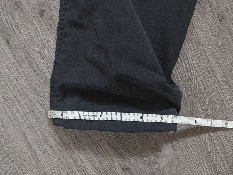 May include: A pair of dark navy blue trousers with a straight hem. A white measuring tape is positioned across the bottom of the pant leg, showing a width measurement of approximately 25.4 cm from the folded edge to the side seam.