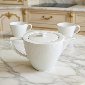 May include: A white porcelain teapot with a subtle vertical ribbed texture, a matching lid with a small knob handle, and a wide spout. Two matching white porcelain teacups with similar ribbed detailing are positioned behind the teapot.