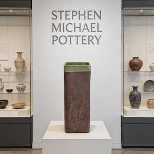 May include: A tall, rectangular brown ceramic vase with a green rim and fern leaf design. The vase is displayed on a white pedestal in a gallery setting. The text "STEPHEN MICHAEL POTTERY" is visible on the wall.