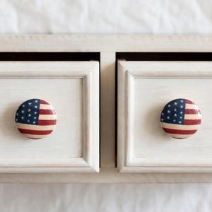 May include: Close-up of two white painted drawer fronts with round knobs. Each knob features the American flag design with red and white stripes and a blue field of white stars. The drawers are slightly ajar, revealing the interior.