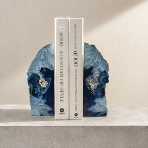 May include: Two blue agate bookends with crystalline centers supporting two books titled AD at 100 A Century of Style and Architectural Digest at 100 The World's Most Beautiful Homes. The bookends feature natural jagged edges and deep blue hues.