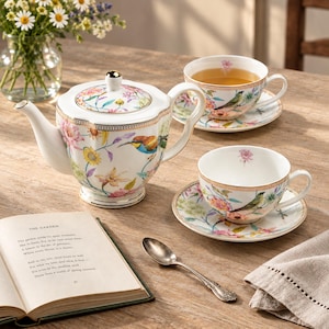 May include: A white porcelain tea set featuring a teapot and two teacups with saucers, decorated with colorful floral patterns and small birds. The set includes gold-toned rims and a silver spoon, displayed alongside an open book titled The Garden.