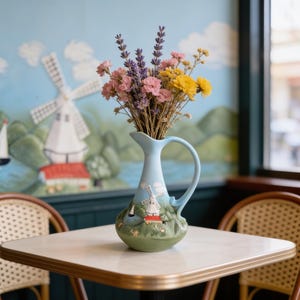 May include: A light blue ceramic pitcher with a handle, decorated with a windmill scene, holds a bouquet of dried flowers. The flowers include pink, yellow, and purple blooms. The pitcher sits on a white table.