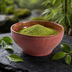 May include: A rustic, reddish-brown ceramic bowl filled with vibrant green matcha powder. Fresh green tea leaves are scattered around the bowl on a dark, textured surface. The background features blurred greenery, suggesting a natural setting.