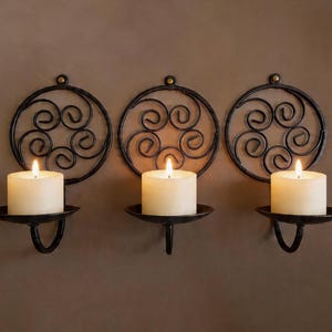 May include: Three black metal wall sconces with lit ivory candles. Each sconce features a circular design with swirling patterns and a small, round candle holder. The candles are cylindrical and provide a warm glow.