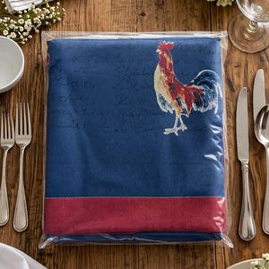 May include: A blue tablecloth with a rooster illustration in red, white, and blue, packaged in clear plastic. The tablecloth has a red stripe at the bottom and script writing. The image is on a wooden table with silverware and a bowl.