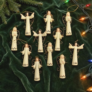 May include: A collection of ten cream-colored angel ornaments, each featuring a textured gown with a small holly leaf and berry detail at the hem. The figures have gold-toned wire wings and halos, with various arm positions including outstretched and folded.