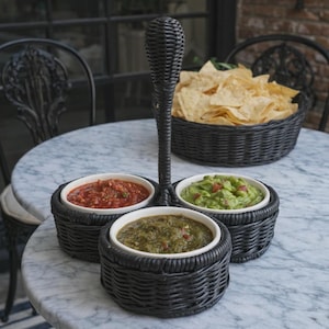 May include: A black woven serving tray with three white ceramic bowls filled with salsa, guacamole, and green salsa. A basket of tortilla chips sits in the background. The tray has a tall handle.