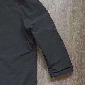 May include: A solid black long-sleeved jacket featuring a smooth, matte fabric finish. The garment displays a clean, structured shoulder seam and a simple, straight sleeve design with a plain cuff, showcasing a uniform dark colour throughout.