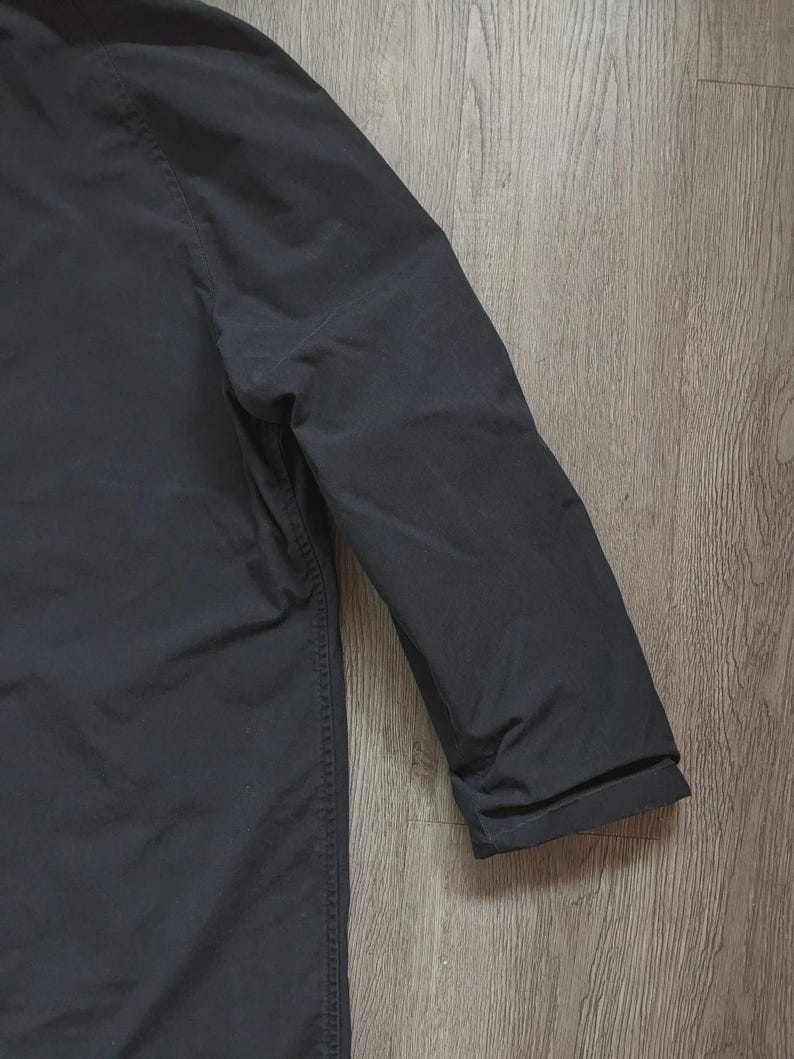 May include: A solid black long-sleeved jacket featuring a smooth, matte fabric finish. The garment displays a clean, structured shoulder seam and a simple, straight sleeve design with a plain cuff, showcasing a uniform dark colour throughout.