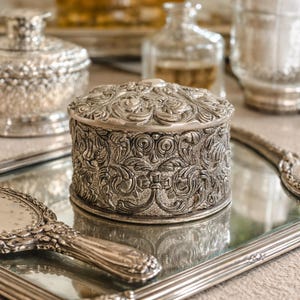 May include: A silver-toned, ornate jewelry box with a detailed floral design on the lid and sides. The box sits on a mirrored tray, alongside a matching hand mirror. Other silver items are visible in the background, suggesting a vintage or antique theme.