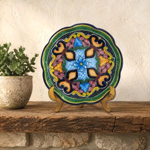 May include: A round ceramic plate with a scalloped edge, featuring a vibrant, symmetrical pattern of blue, yellow, pink, and green floral and scroll motifs. The plate is displayed on a gold-toned metal stand, showcasing intricate hand-painted details.