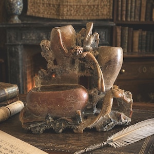 May include: A detailed, antique brown stone sculpture with a central bowl, two vases, and carved bird and floral details. The sculpture sits on a wooden desk with a quill pen and paper, suggesting a writing or study theme.