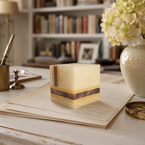 May include: A square decorative box made of smooth, cream-colored stone featuring horizontal and vertical inlaid bands of dark brown wood and light yellow wood. The box has a clean, geometric design with sharp edges and a polished, matte finish.