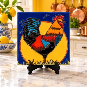 May include: A square ceramic tile featuring a colorful rooster illustration against a bright yellow sun and deep blue background. The rooster has red, orange, and black plumage. The tile is supported by a black plastic display stand.