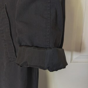 May include: A black long-sleeved coat featuring a structured fabric exterior and a contrasting smooth, dark black lining visible at the folded cuff. The garment displays visible vertical stitching along the sleeve and a clean, minimalist design.