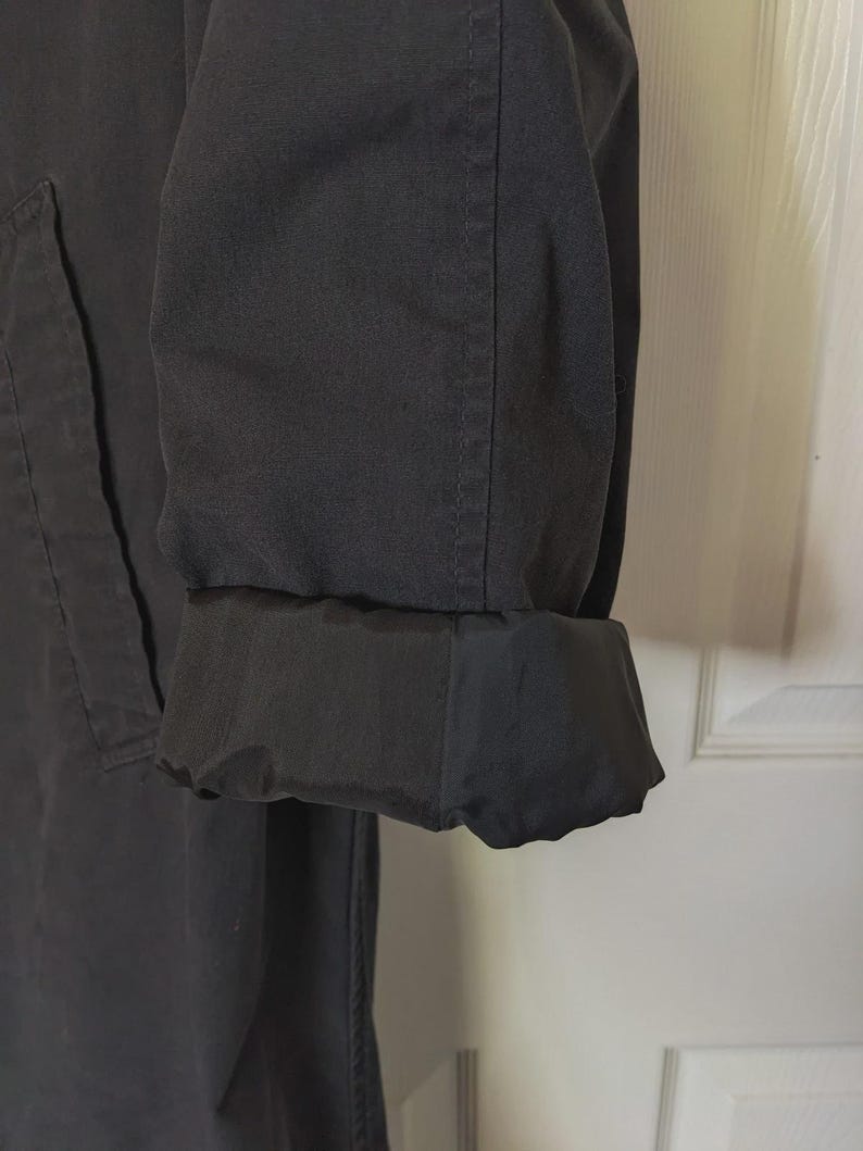 May include: A black long-sleeved coat featuring a structured fabric exterior and a contrasting smooth, dark black lining visible at the folded cuff. The garment displays visible vertical stitching along the sleeve and a clean, minimalist design.