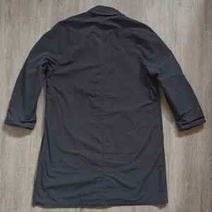 May include: A solid dark charcoal grey long coat featuring a classic collar, long sleeves with cuffs, and a prominent vertical seam running down the centre of the back. The garment is constructed from a smooth, matte fabric with visible stitching.