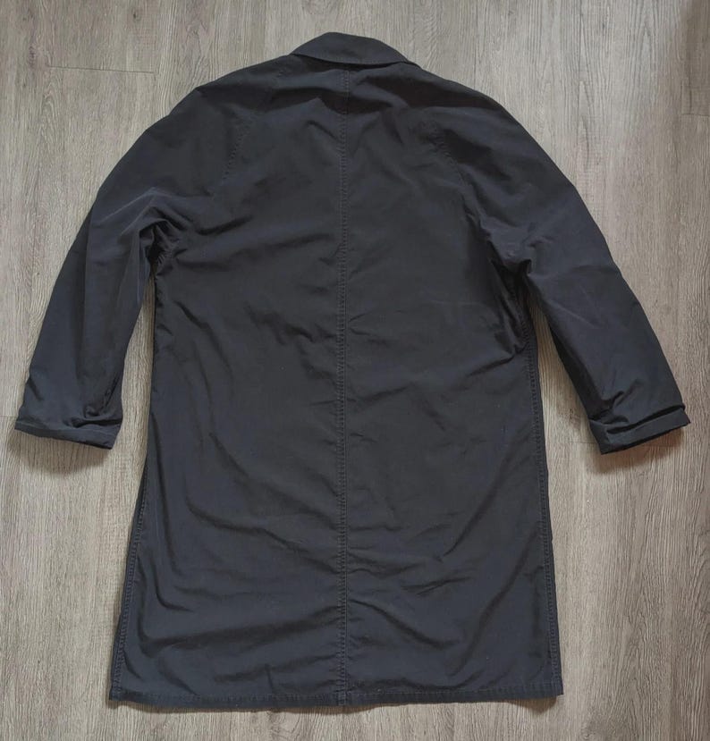 May include: A solid dark charcoal grey long coat featuring a classic collar, long sleeves with cuffs, and a prominent vertical seam running down the centre of the back. The garment is constructed from a smooth, matte fabric with visible stitching.