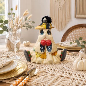 May include: A cream-colored ghost figurine wearing a yellow and blue baseball cap with a small black creature on top. The ghost has large blue eyes, red gloves, and a dress featuring blue polka-dot sleeves and yellow patches, sitting with dark shoes.