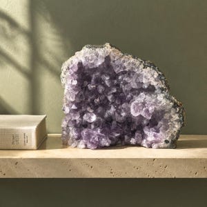 May include: A large amethyst geode featuring deep purple, crystalline structures within a rough, greyish-brown rock exterior. The cluster displays numerous pointed, translucent violet points that catch the light, creating a textured, jagged surface.
