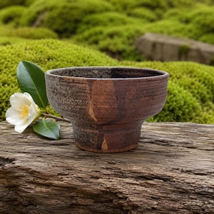 May include: A small, wide-rimmed ceramic bowl with a textured, dark brown and charcoal-grey mottled finish. The bowl features a tapered base and a rough, earthy surface appearance. A single white camellia flower with green leaves sits beside it.