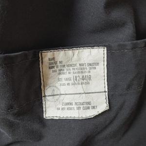 May include: A black garment label with white text reading: NAME, SERVICE NO, NAME OF ITEM: RAINCOAT, BASE FABRIC: 50% POLYESTER/50% COTTON, CONTRACT NO: DLA100-86-C-0736, SIZE: LARGE (42-44)R, STOCK NO: 8405-00-304-3566, CLEANING INSTRUCTIONS: FOR BEST RESULTS, DRY CLEAN ONLY.
