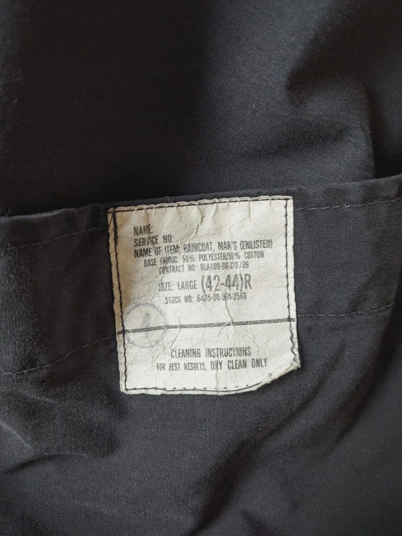 May include: A black garment label with white text reading: NAME, SERVICE NO, NAME OF ITEM: RAINCOAT, BASE FABRIC: 50% POLYESTER/50% COTTON, CONTRACT NO: DLA100-86-C-0736, SIZE: LARGE (42-44)R, STOCK NO: 8405-00-304-3566, CLEANING INSTRUCTIONS: FOR BEST RESULTS, DRY CLEAN ONLY.