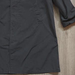 May include: A long, dark charcoal grey coat featuring a concealed front button placket, a structured collar, and a single visible slanted welt pocket on the side. The garment has long sleeves and a straight hemline, crafted from a smooth, matte fabric.