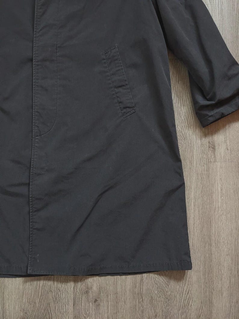 May include: A long, dark charcoal grey coat featuring a concealed front button placket, a structured collar, and a single visible slanted welt pocket on the side. The garment has long sleeves and a straight hemline, crafted from a smooth, matte fabric.