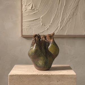 May include: A hand-formed ceramic vase with an irregular, organic shape. The surface features a mottled glaze in shades of moss green, olive, and deep brown, with a rough, textured finish and a pinched, uneven rim at the top of the vessel.