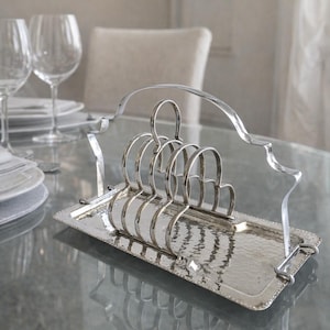 May include: A silver-toned metal toast rack with a hammered texture, holding several arched slots. The rack sits on a rectangular tray with a decorative edge and a curved handle. The setting includes glassware and tableware, suggesting a dining theme.