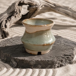 May include: A small ceramic pot with a flared rim and a tapered base, featuring a glossy light green glaze with visible crackle patterns and earthy brown accents along the rim and base, showcasing a distinct hand-thrown pottery aesthetic.