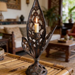 May include: Ornate, dark brown metal candle holder with a lit, cream-colored candle. The candle holder has a leaf-like design with floral cutouts. The base is round with a geometric pattern. The candle holder is on a bamboo table.