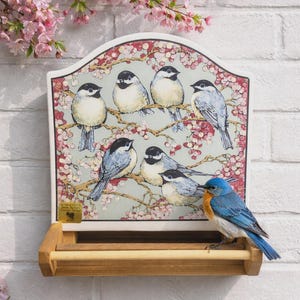 May include: A decorative bird feeder with a painted illustration of several birds perched on a flowering branch. The feeder has a wooden base and a blue bird is perched on the feeder's edge. The background is a white brick wall.