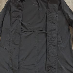 May include: A black long-sleeve coat featuring a button-down front closure with a visible placket. The interior lining displays the printed text RCS 9170 115. The garment has a structured collar and a smooth, matte fabric finish throughout the design.