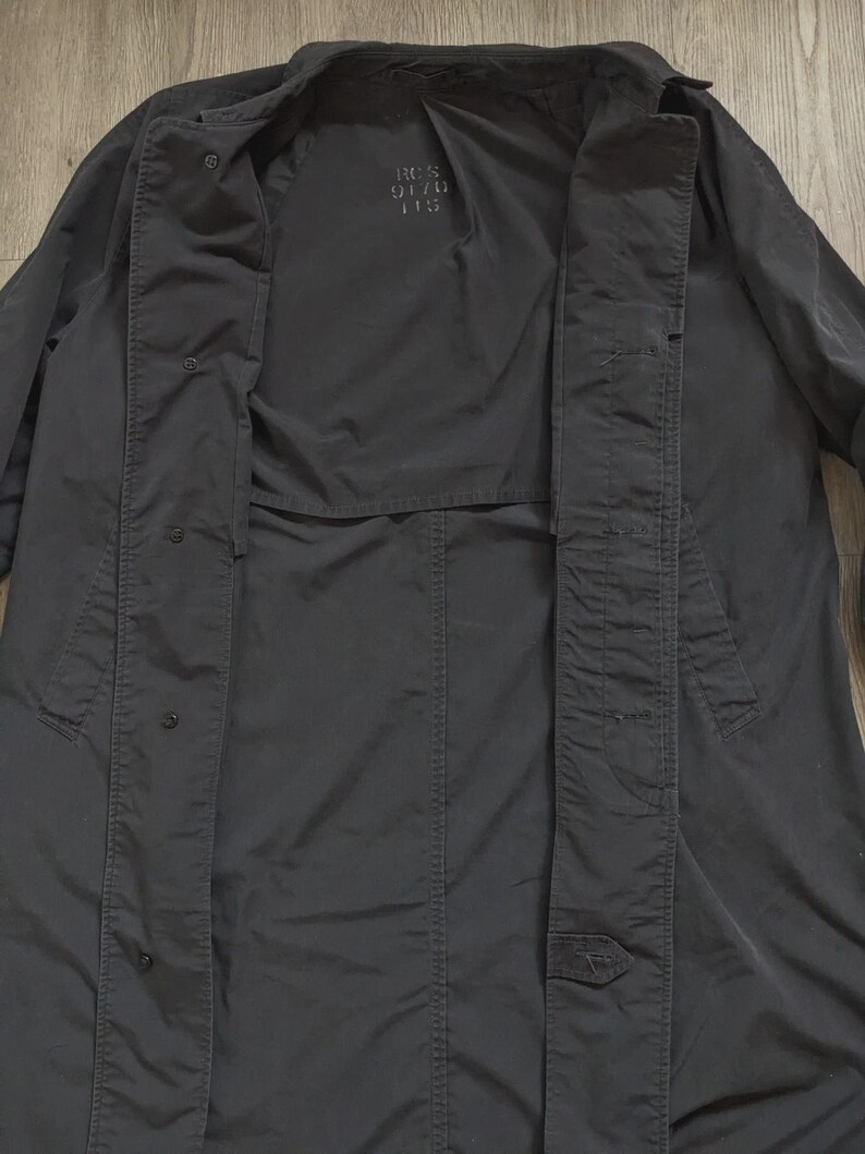 May include: A black long-sleeve coat featuring a button-down front closure with a visible placket. The interior lining displays the printed text RCS 9170 115. The garment has a structured collar and a smooth, matte fabric finish throughout the design.