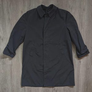 May include: A solid dark charcoal grey raincoat featuring a classic pointed collar, long sleeves, and a concealed front button placket. The coat has two vertical side pockets and a straight hemline, providing a clean and functional outer layer.