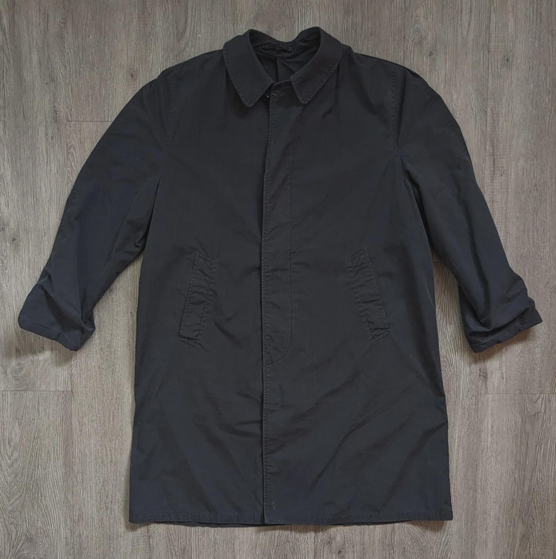 May include: A solid dark charcoal grey raincoat featuring a classic pointed collar, long sleeves, and a concealed front button placket. The coat has two vertical side pockets and a straight hemline, providing a clean and functional outer layer.