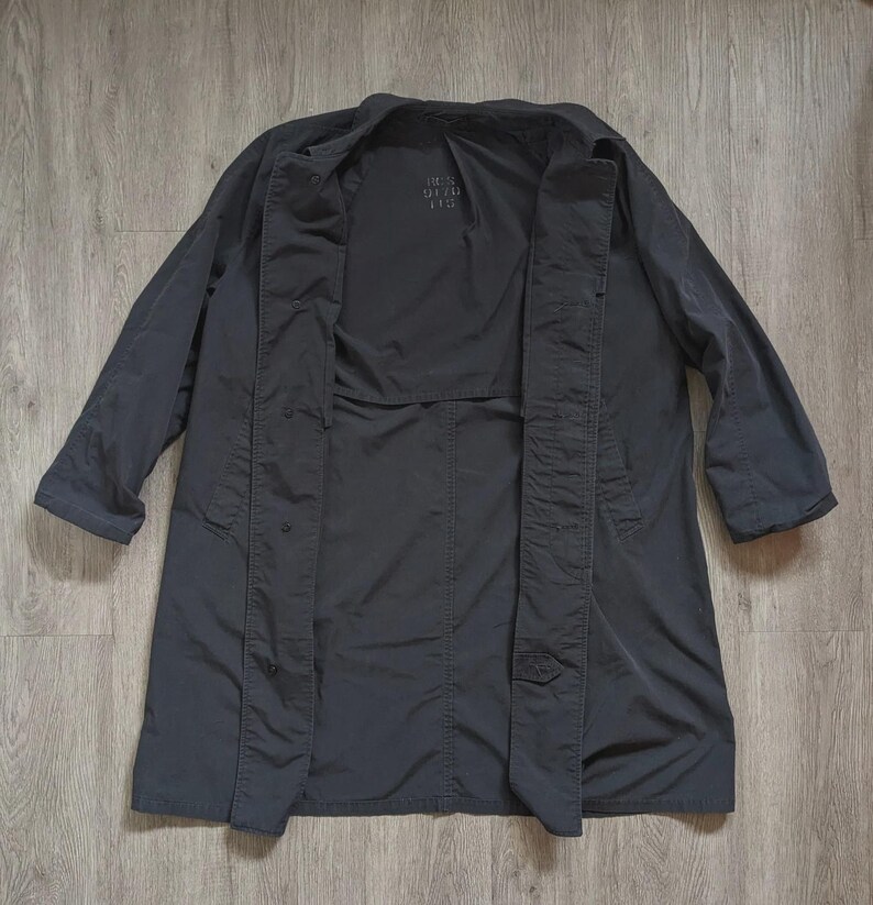 May include: A dark charcoal grey trench coat featuring a wide collar, long sleeves, and a front button closure. The interior lining displays the text "BEN DITO US" printed in a subtle font. The coat has a structured silhouette with visible seams.